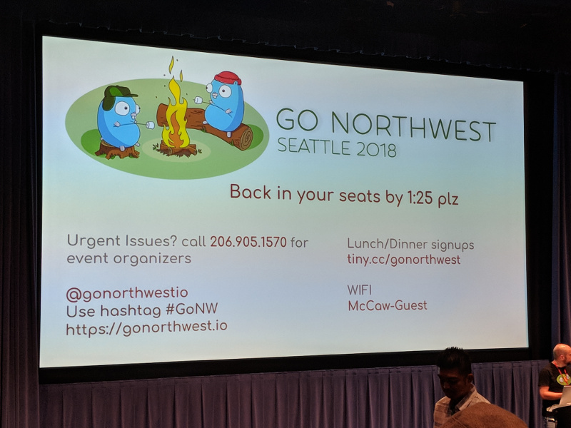 Projector screen showing a slide describing a break at the conference and displaying the Twitter handle @gornorthwestio, the hashtag #GoNW, the website https://gonorthwest.io, emergency contact information for the organizers, lunch/dinner sign ups, and the WiFi SSID